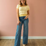 Woman wearing a yellow striped shirt and blue jeans standing against a pink wall.
