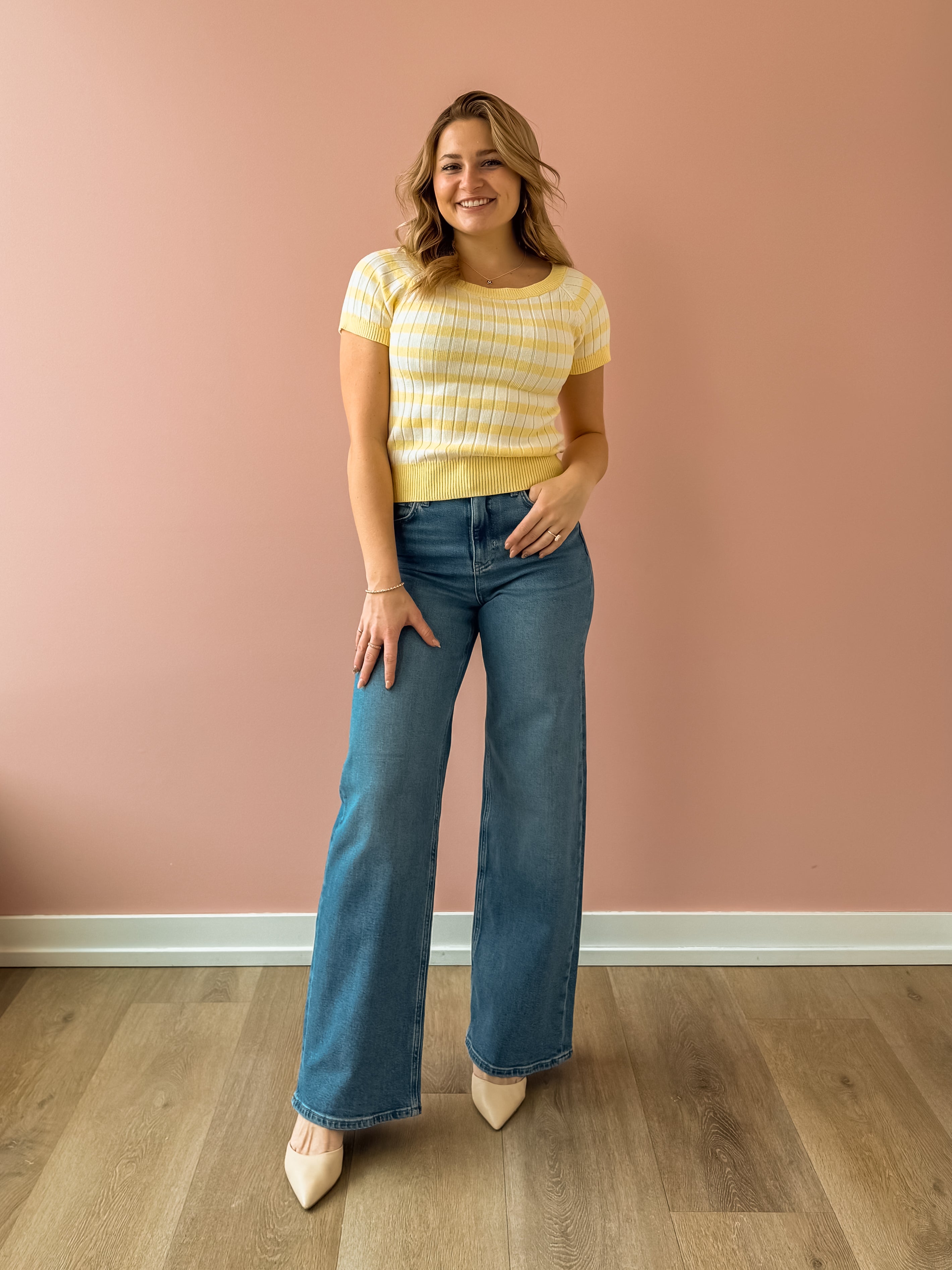 Woman wearing a yellow striped shirt and blue jeans standing against a pink wall.