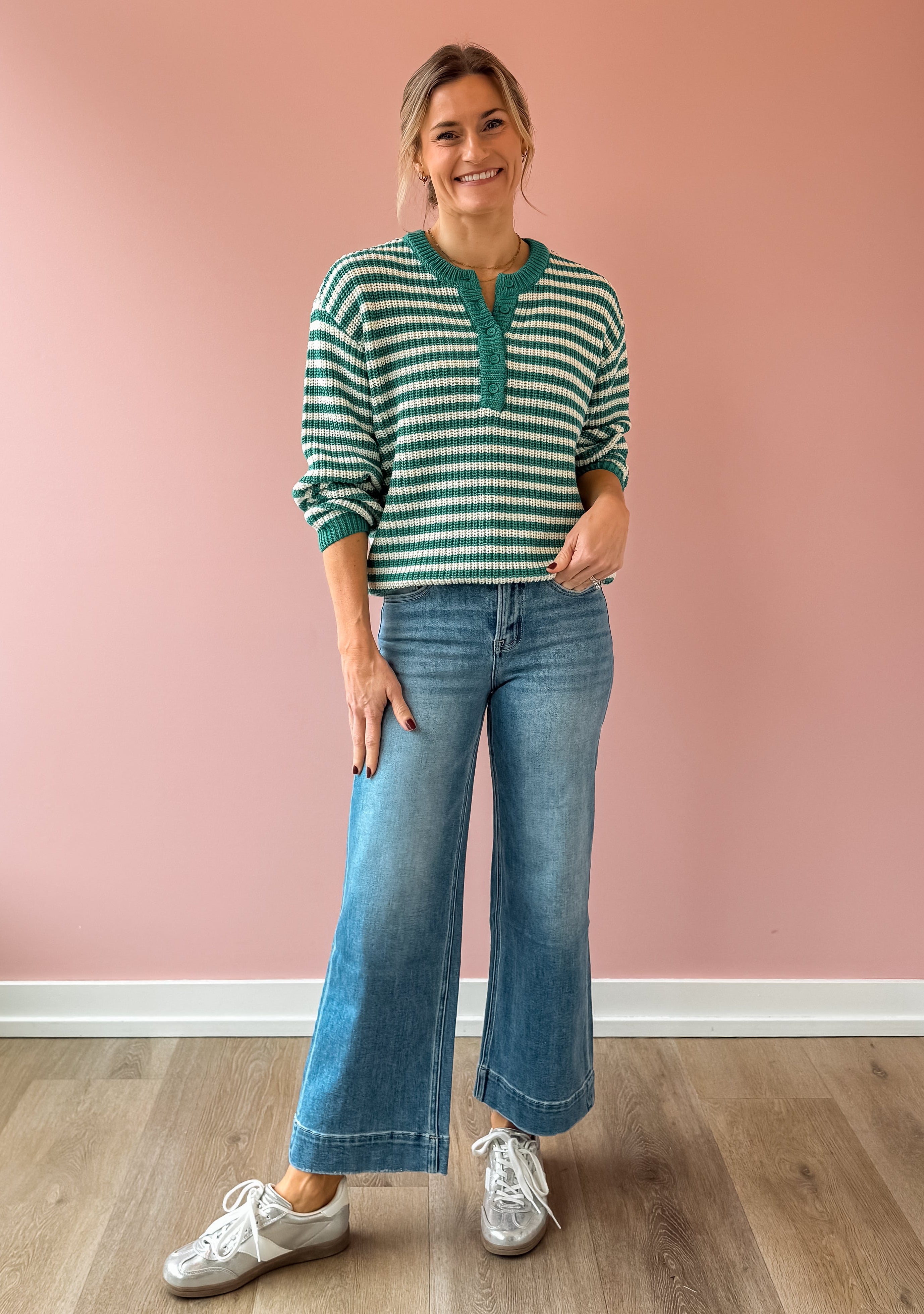 Woman wearing a green and white striped sweater and blue jeans standing against a pink wall.
