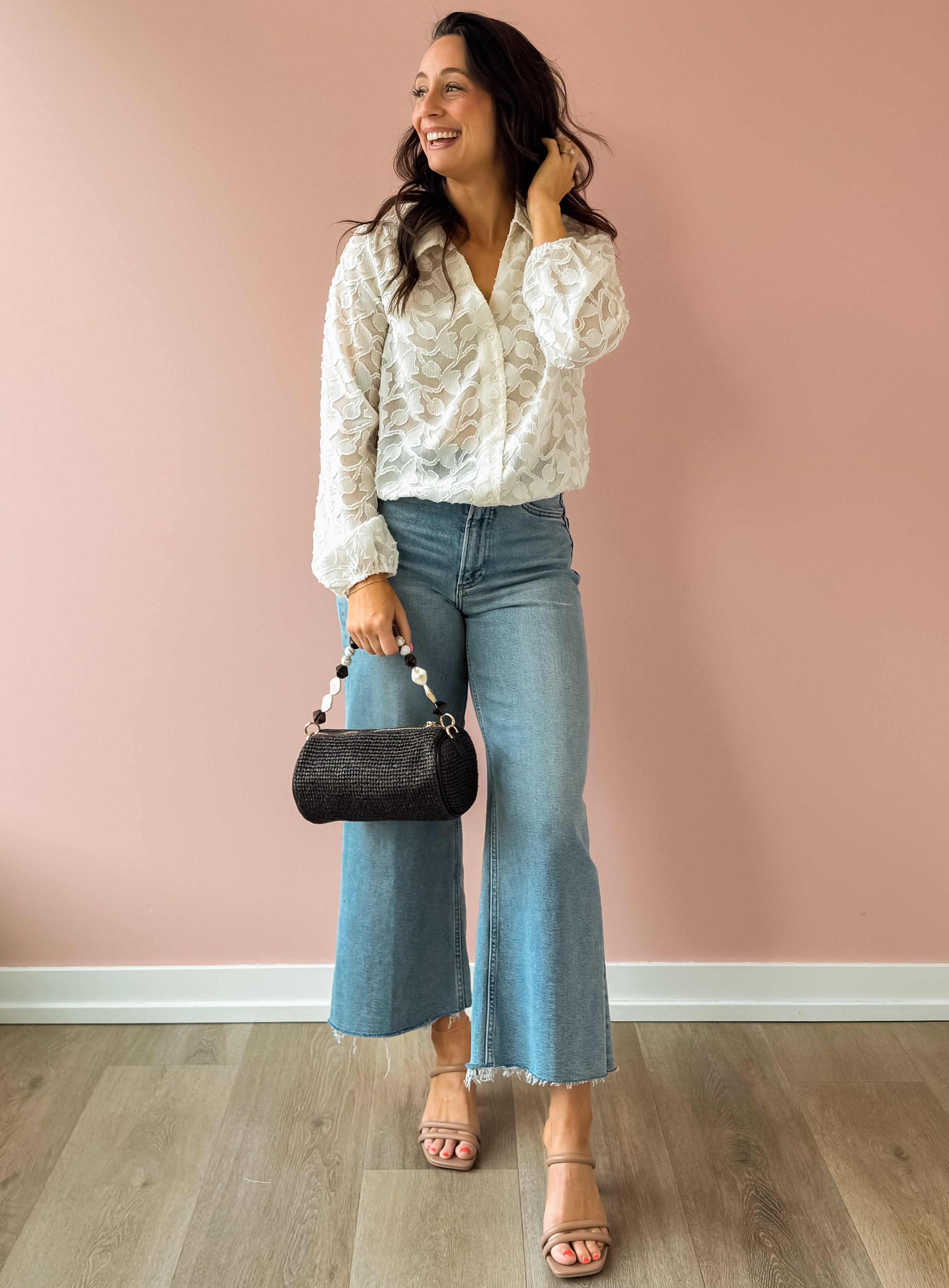 Woman wearing a white blouse and blue jeans, holding a black handbag against a pink wall.